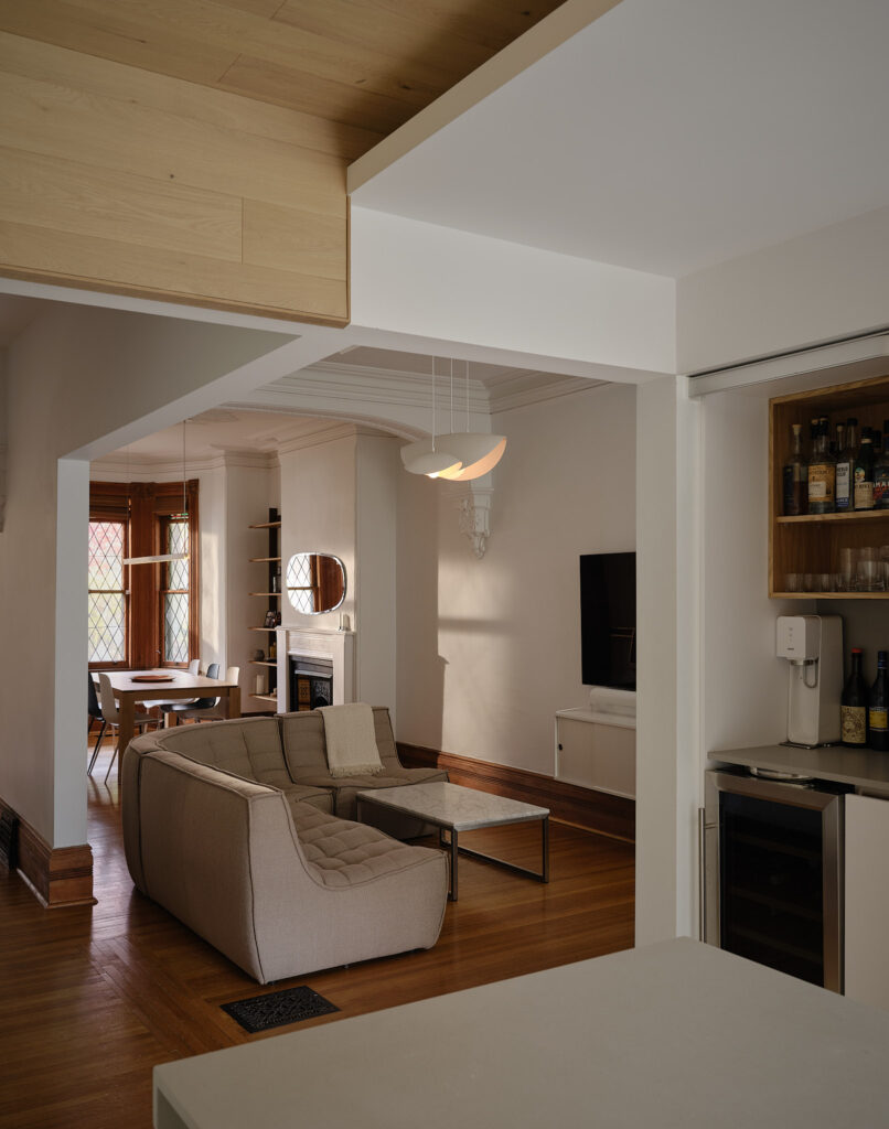 Looking from the kitchen to the living and dining rooms. The bar can be seen on the right. Sunlight shines in through the front windows onto the dining table and fireplace.